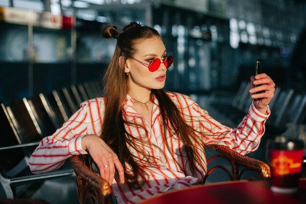 かわいいファッションモデルは、夕日の光の中でカフェに座っている彼女のスマートフォンで動作します