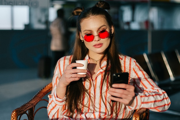 Il modello di moda grazioso funziona con il suo smartphone che si siede al caffè nei raggi del tramonto