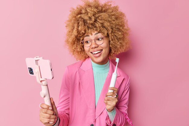 Pretty female blogger shows her morning routine during blog live stream brushes teeth with modern electric toothbrush wears formal jacket isolated over pink background takes photos on cellular