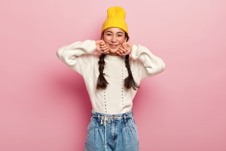 無料Photo ファッショナブルな黄色の帽子、白いセーター、ジーンズに身を包んだきれいな女性は、ピンクの壁に隔離された、カメラの熱狂的で魅力的な外観、嬉しい表情をしています