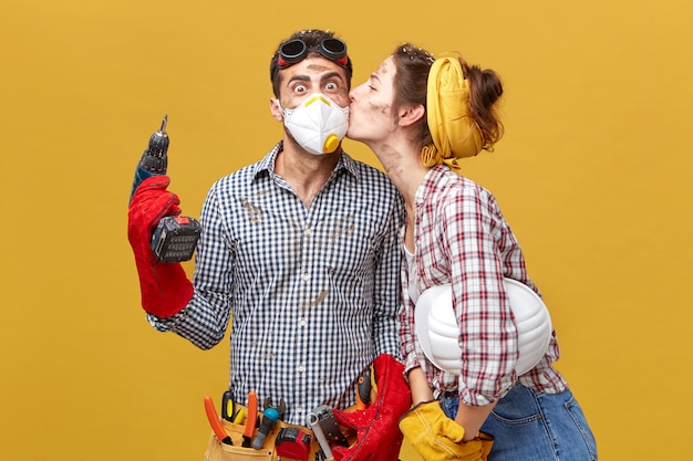 Free photo pretty female kissing her husband in cheek being thankful to him for repairing her wardrobe. surprised male worker in mask holding drilling machine being glad to receive kiss from his girlfriend