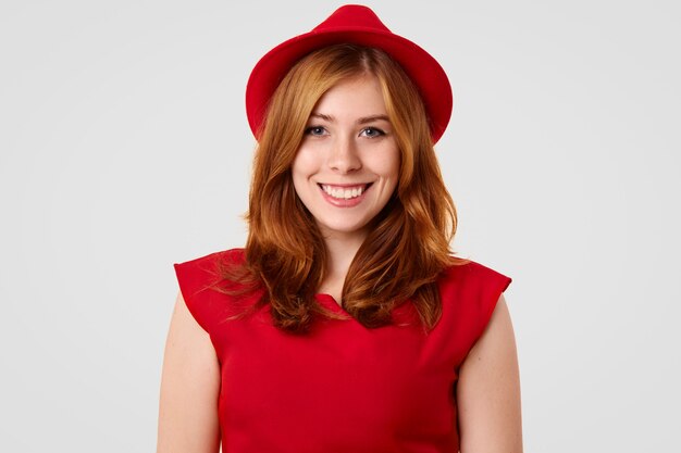 Pretty female model with positive smile dressed in elegant red hat and blouse, going to have date with boyfriend