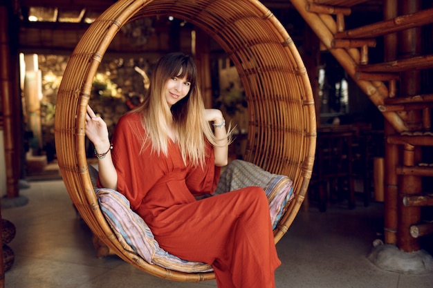 Pretty female siting in hanging bamboo stair on open-air veranda of wooden bungalow