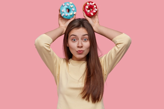 Foto gratuita la giovane donna castana abbastanza divertente tiene due ciambelle deliziose, vestite in vestiti gialli, modelli sopra lo spazio rosa