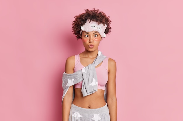 Free photo pretty funny curly haired afro american woman keeps lips folded crosses eyes and makes funny grimace dressed in nightwear poses against pink wall