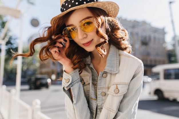 黄色いサングラスにふざけて触れている生姜少女。街で時間を過ごす帽子をかぶった愛らしい赤毛の女性の屋外写真。