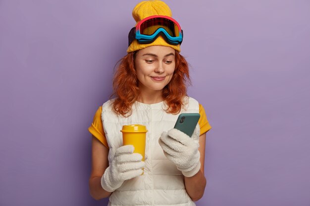 Pretty ginger woman rests after winter sports, checks email box, holds takeaway cup of coffee, wears snowboarding goggles, isolated over purple wall