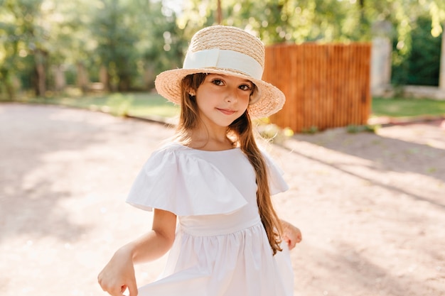 Pretty girl in big straw hat playing with her white dress while posing in park with wooden fence. Portrait of lovely female kid wears boater decorated with ribbon dancing on the road.