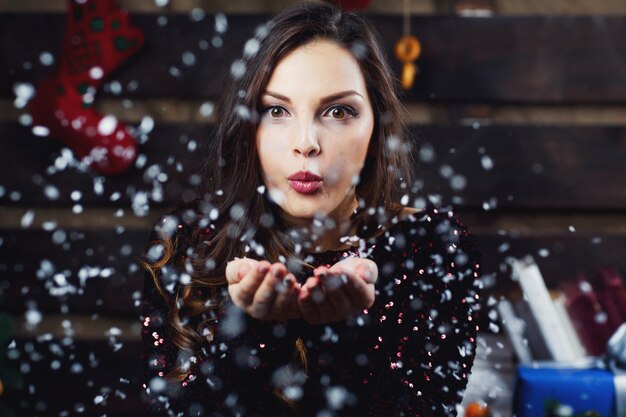 Красивая девушка сносит снег с ладоней, стоящих в комнате, приготовленной к рождественским праздникам