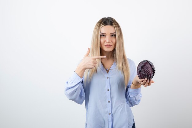 Pretty girl in blue outfit pointing at purple cabbage on white. 