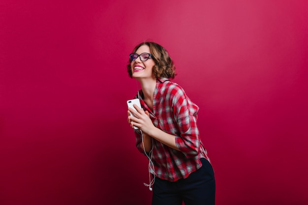 Pretty girl in checkered shirt laughing with eyes closed on claret wall. Indoor photo of emotional curly short-haired woman in glasses holding phone.