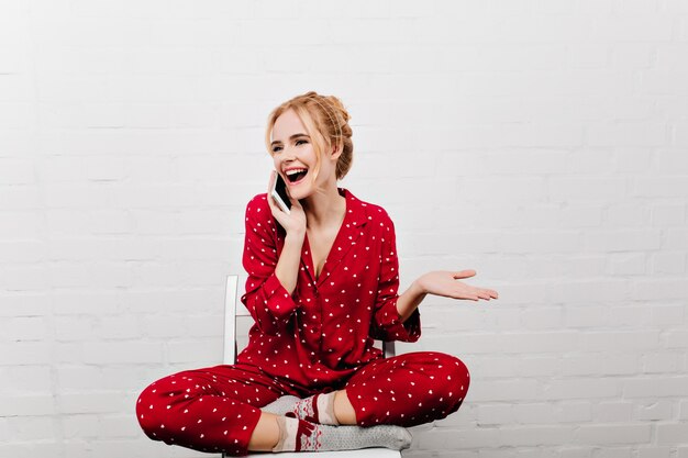 朝の電話で話しているかわいい靴下のかわいい女の子。白い壁に足を組んで座っている赤いパジャマを着たポジティブな白人女性。