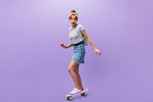 Pretty girl in denim skirt rides longboard. Wonderful cool young woman in grey trendy shirt and white sneakers posing on isolated background.