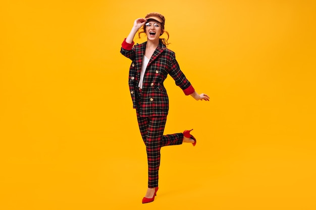 Pretty girl in eyeglasses, cap and suit jumps on orange wall