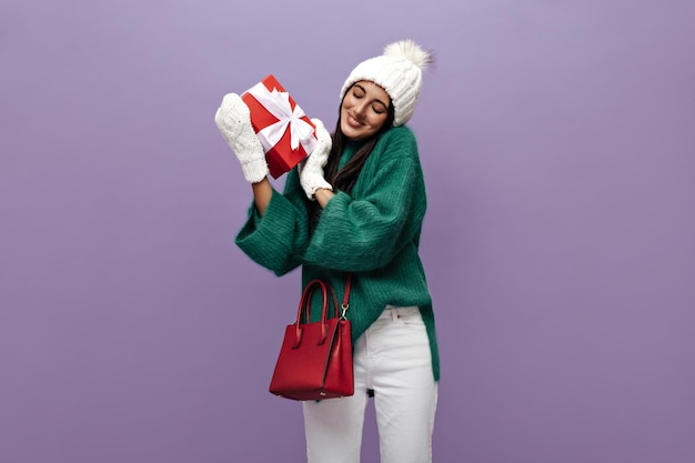 Pretty girl in green sweater and white pants poses with red gift box Charming lady in woolen hat and knitted gloves smiles on purple background