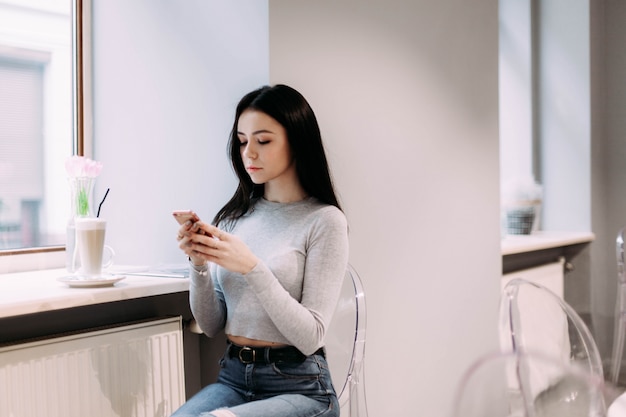 Pretty girl in grey shirt reads something in her phone sitting in the cafe