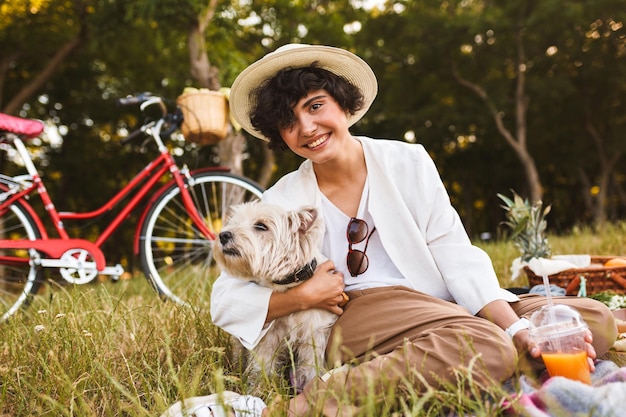 Bella ragazza con cappello che abbraccia un piccolo cane carino che guarda gioiosamente nella fotocamera trascorrendo del tempo a fare un picnic nel parco con una bicicletta rossa sullo sfondo