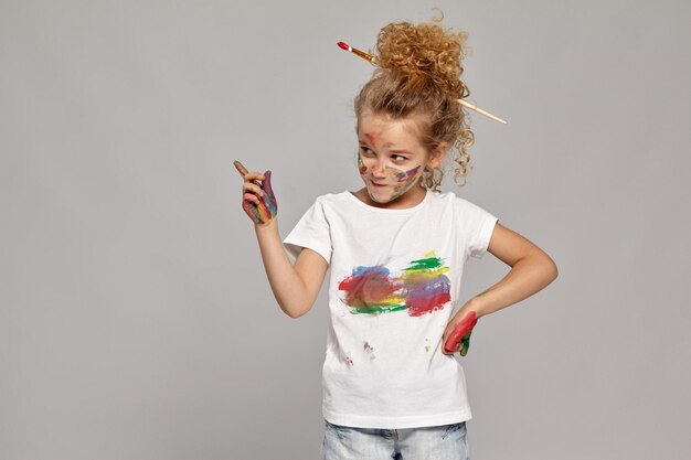 Pretty girl having a brush in her lovely haircut, wearing in a white smeared t-shirt. She is posing with a painted hands and cheeks, looking angry and pointing on someone, on a gray background.