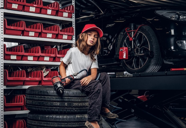 Pretty girl is posing for photographer while sitting at dark auto service holding pneumatic drill.