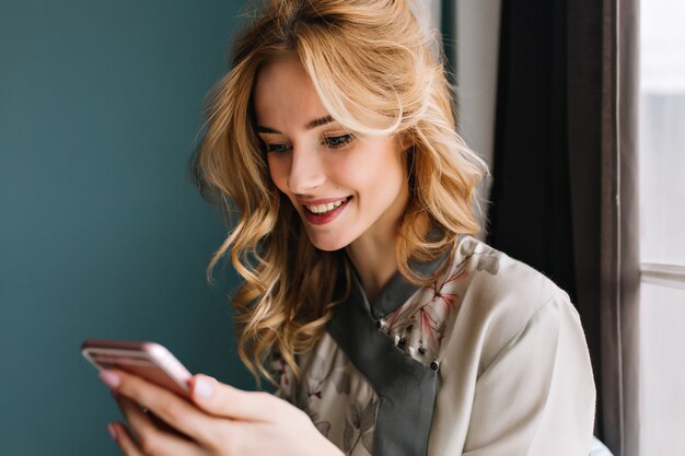 Pretty girl looking on her pink phone and smiling, wearing silk pajama, sitting on window sill. She has beautiful wavy blonde hair. Room with turquoise wall.