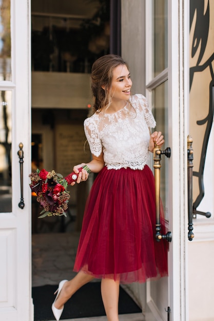Pretty girl in marsala tulle skirt on street. She holds flowers and smiling to side