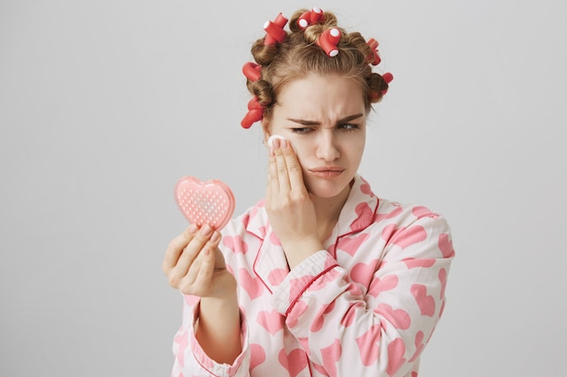 Free photo pretty girl in nighwear and hair curlers, looking mirror and wipe-off makeup up cotton pad