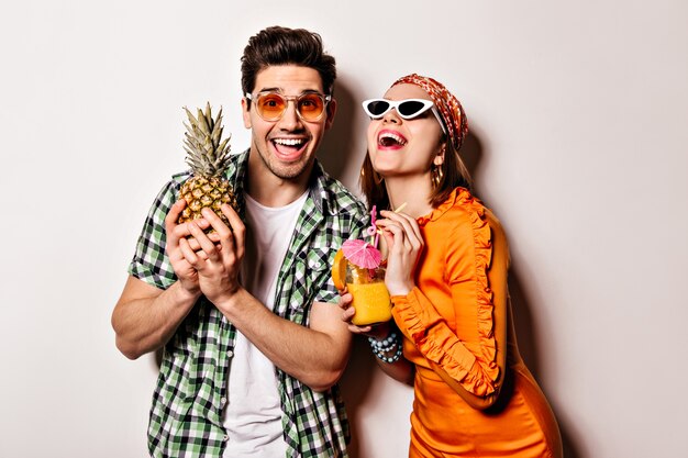 Pretty girl in orange dress and guy in green shirt and sunglasses are laughing and posing with pineapple and cocktail.