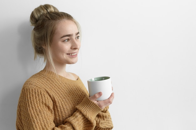 Free photo pretty girl in oversize sweater warming up on cold autumn day, having coffee, holding large mug and smiling joyfully. attractive young female with hair knot relaxing at home with cup of tea