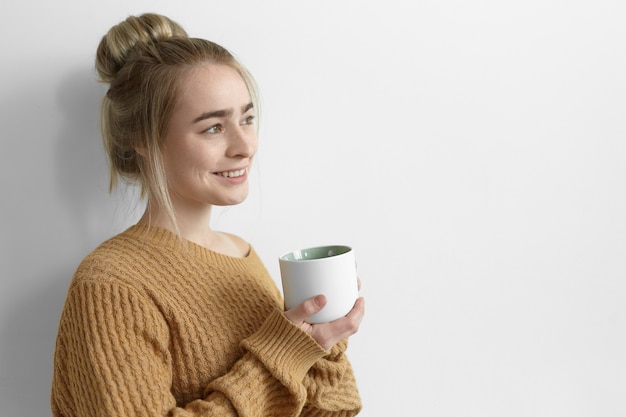 Pretty girl in oversize sweater warming up on cold autumn day, having coffee, holding large mug and smiling joyfully. Attractive young female with hair knot relaxing at home with cup of tea