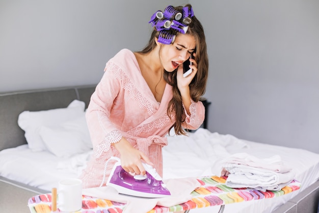 Free photo pretty girl in pink bathrobe and curler ironing clothes and speaking on phone at home. she looks astonished and busy.