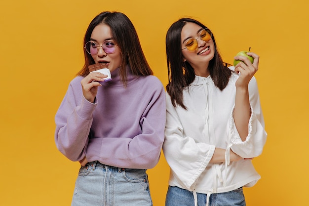 Pretty girl in purple sweater bites milk chocolate bar Young woman in white hoodie looks at green apple and smiles Portrait of charming Asian women pose on orange background