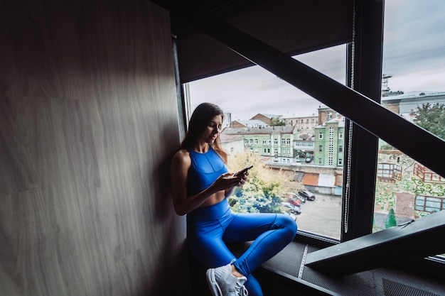 Pretty girl sitting on the window sill with smartphone in hands.