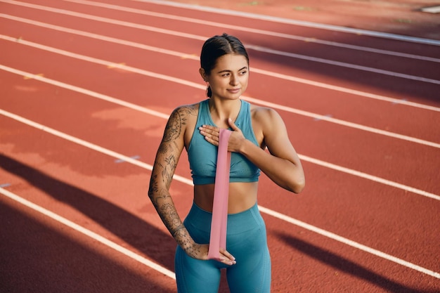 Pretty girl in stylish sportswear dreamily stretching hands with rubber band during workout on city stadium