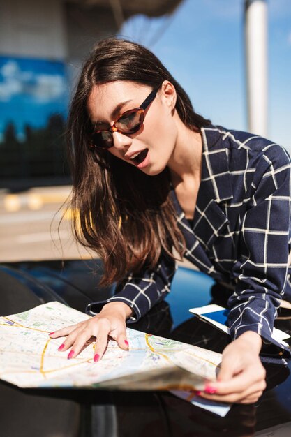 Pretty girl in sunglasses leaning on black car while amazedly looking road on map with airport on background