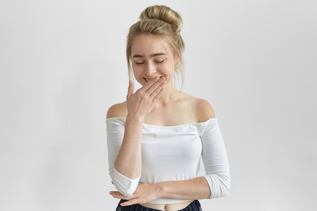 Free photo pretty girl wearing stylish top with open shoulders keeping eyes closed and covering mouth with hand while giggling, listening to funny story. attractive young female with hair knot laughing at joke