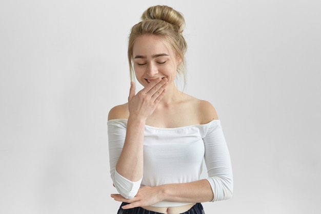 Bella ragazza che indossa un top elegante con le spalle aperte, tenendo gli occhi chiusi e coprendo la bocca con la mano mentre rideva, ascoltando una storia divertente. attraente giovane femmina con nodo di capelli ridendo per scherzo