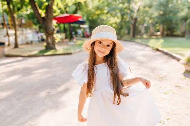 Pretty girl with beautiful big dark eyes posing while having fun in park in summer vacation. Outdoor portrait of funny long-haired child in straw hat standing on the road with surprised smile.
