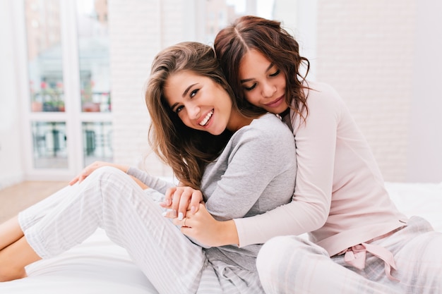 Pretty girl with curly hair is hugging from back other girl smiling . They wear pajamas.