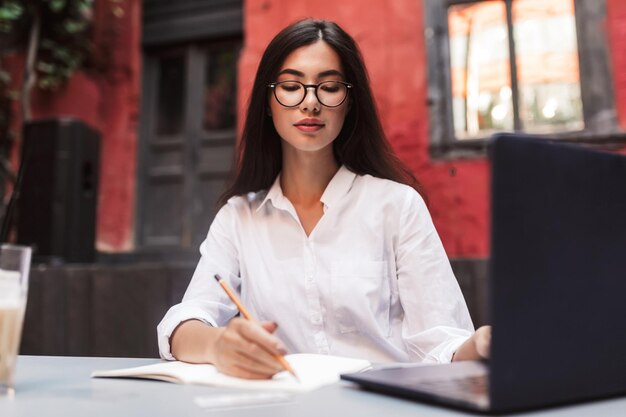 Pretty girl with dark hair in white shirt and eyeglasses dreamily working with laptop and notepad in cozy courtyard of cafe