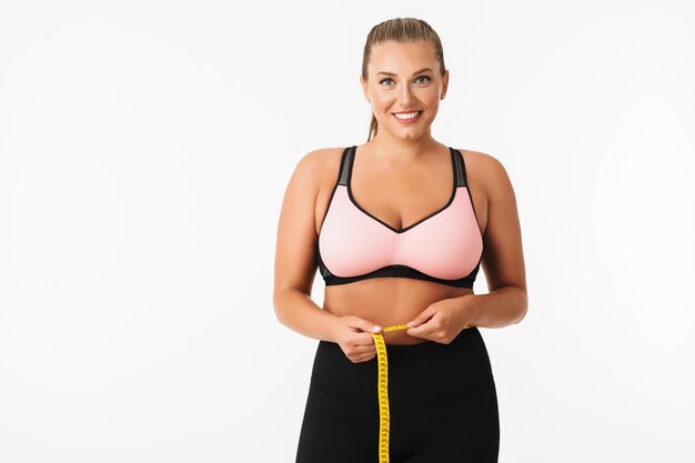 Pretty girl with excess weight in sporty top measuring waist while happily looking in camera over white background