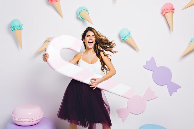 Pretty girl with hair waving wearing violet skirt singing favorite song and holding toy lollipop. Portrait of stylish young woman with eyes closed having fun on party and dancing.