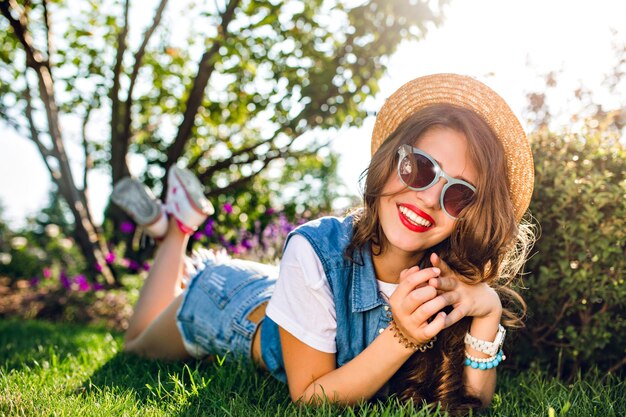 Красивая девушка с длинными вьющимися волосами в шляпе лежит на траве в летнем парке. на солнечном свете. На ней джинсовые куртки, шорты, солнцезащитные очки. Она улыбается в камеру с красными губами.