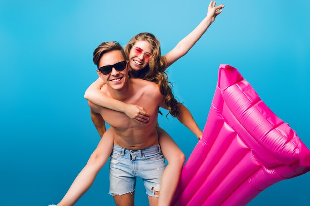 Pretty girl with long curly hair is riding on back of handsome guy with naked torso on blue background in studio. He holds pink mattress and they smile to camera. They have fun together.