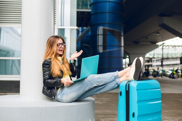 Pretty girl with long hair in black glasses is sitting outside in airport. She wears jeans, black jacket, jellow shoes. She put her legs on suitcase and speaking on laptop. She looks happy.