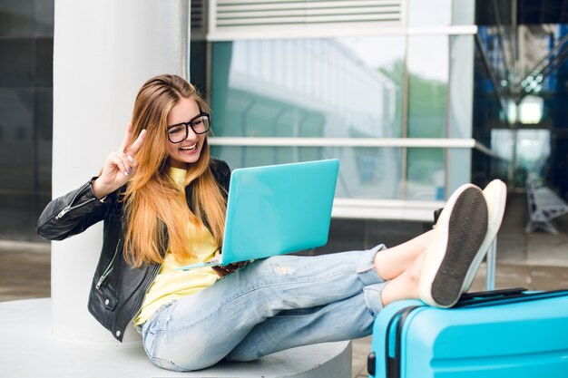 Pretty girl with long hair in black glasses is sitting outside in airport. She wears jeans, black jacket, jellow shoes. She put her legs on suitcase and speaking on laptop.