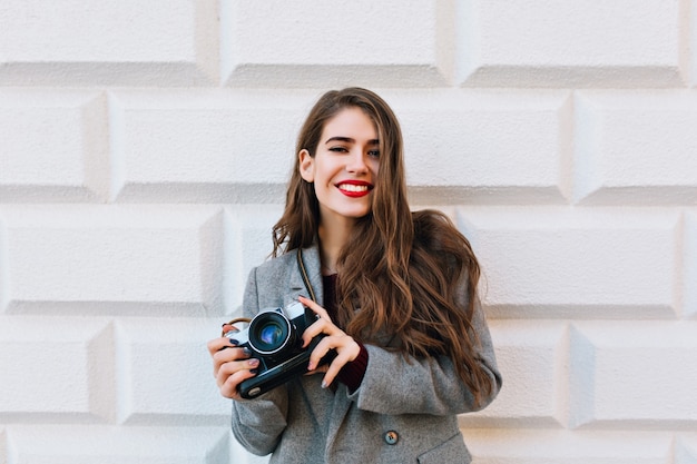 Pretty girl with long hair in grey coat on wall . She holds camera and smiling with red lips.