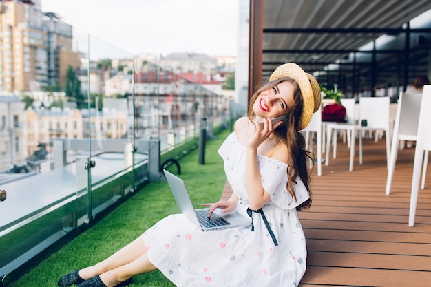 Pretty girl with long hair in hat is sitting on floor on the terrace. She wears a white dress with naked shoulders. She is typing on  laptop and speaking on phone.