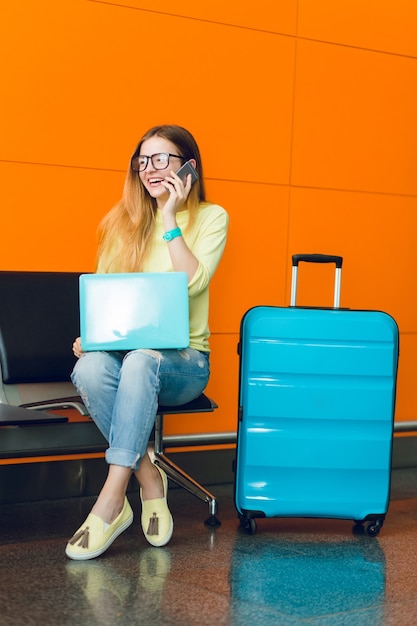 Pretty girl with long hair is sitting on chair on orange background. She wears jeans with jellow sweater. She has laptop and suitcase near. She is speaking on phone.