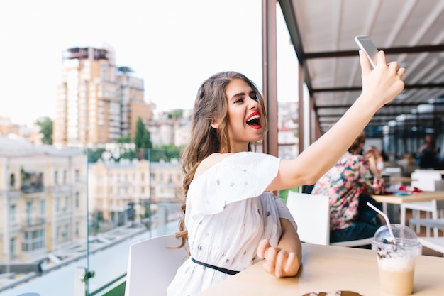 Pretty girl with long hair is sitting at  table on the terrace in cafe . She wears a white dress with bare shoulders and red lipstick . She is making selfie-portrait with phone.,