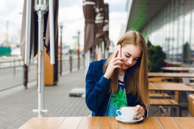 A pretty girl with long hair is sitting at the table on the terrace. She is speaking on phone and holds a cup of coffee.